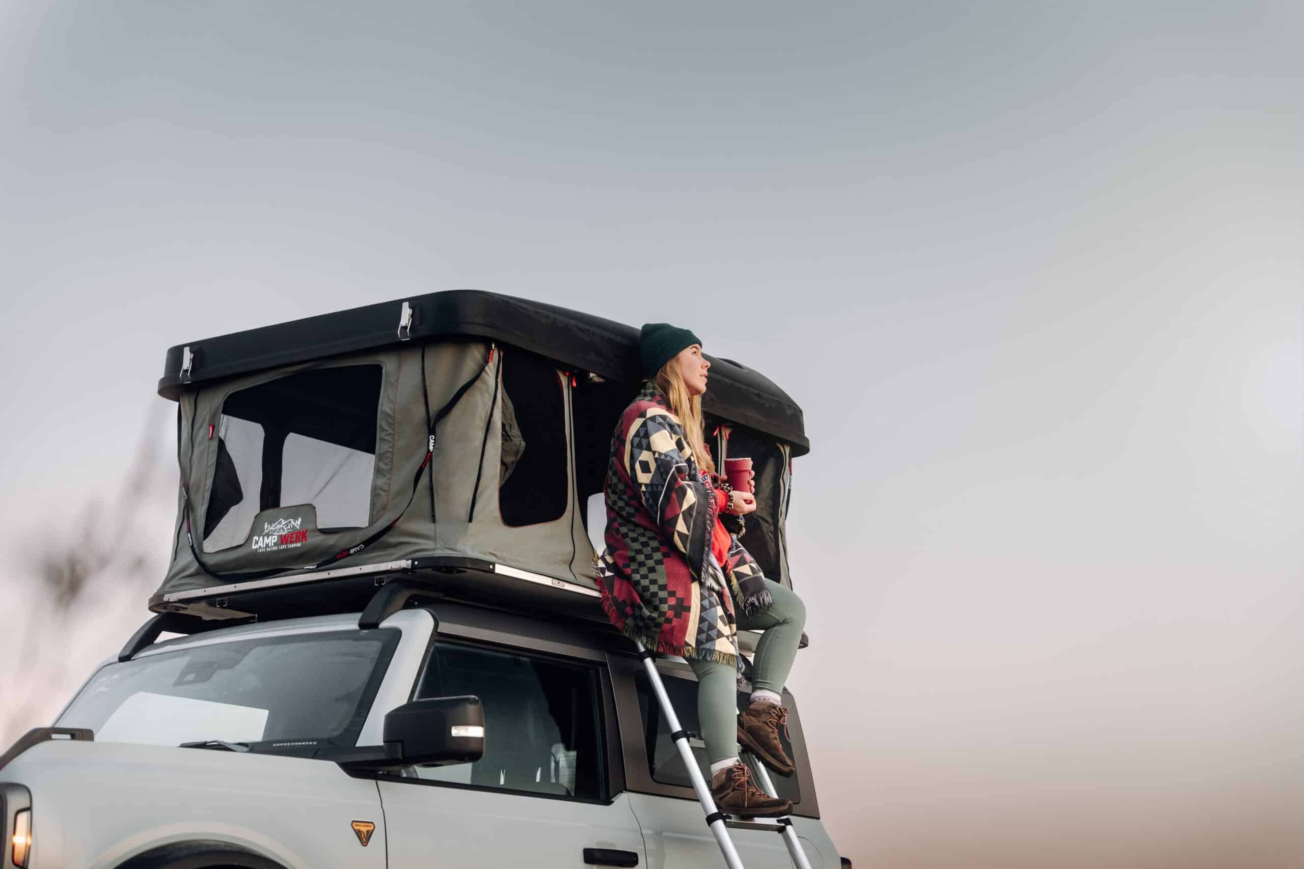 Una donna è seduta all'ingresso della sua tenda da tetto e si gode la natura. Nella Good Vibes c'è abbastanza spazio per questo, perché offre una sorprendente altezza libera. La Good Vibes è la prima tenda da tetto rigida ad apertura verticale di Campwerk.