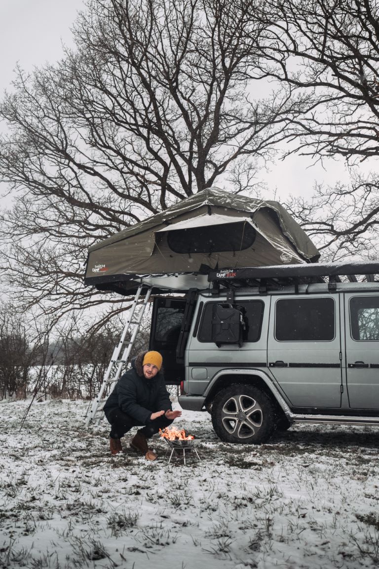 Camperista si scalda le mani al fuoco accanto alla sua auto con tenda da tetto nella neve