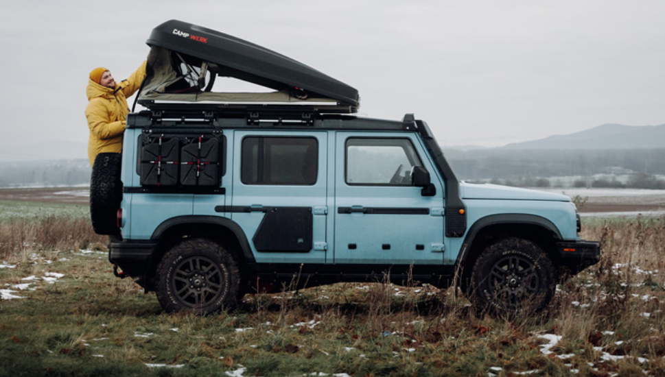 Uomo in giacca antipioggia gialla apre la sua tenda da tetto Good Vibes su un'auto azzurra
