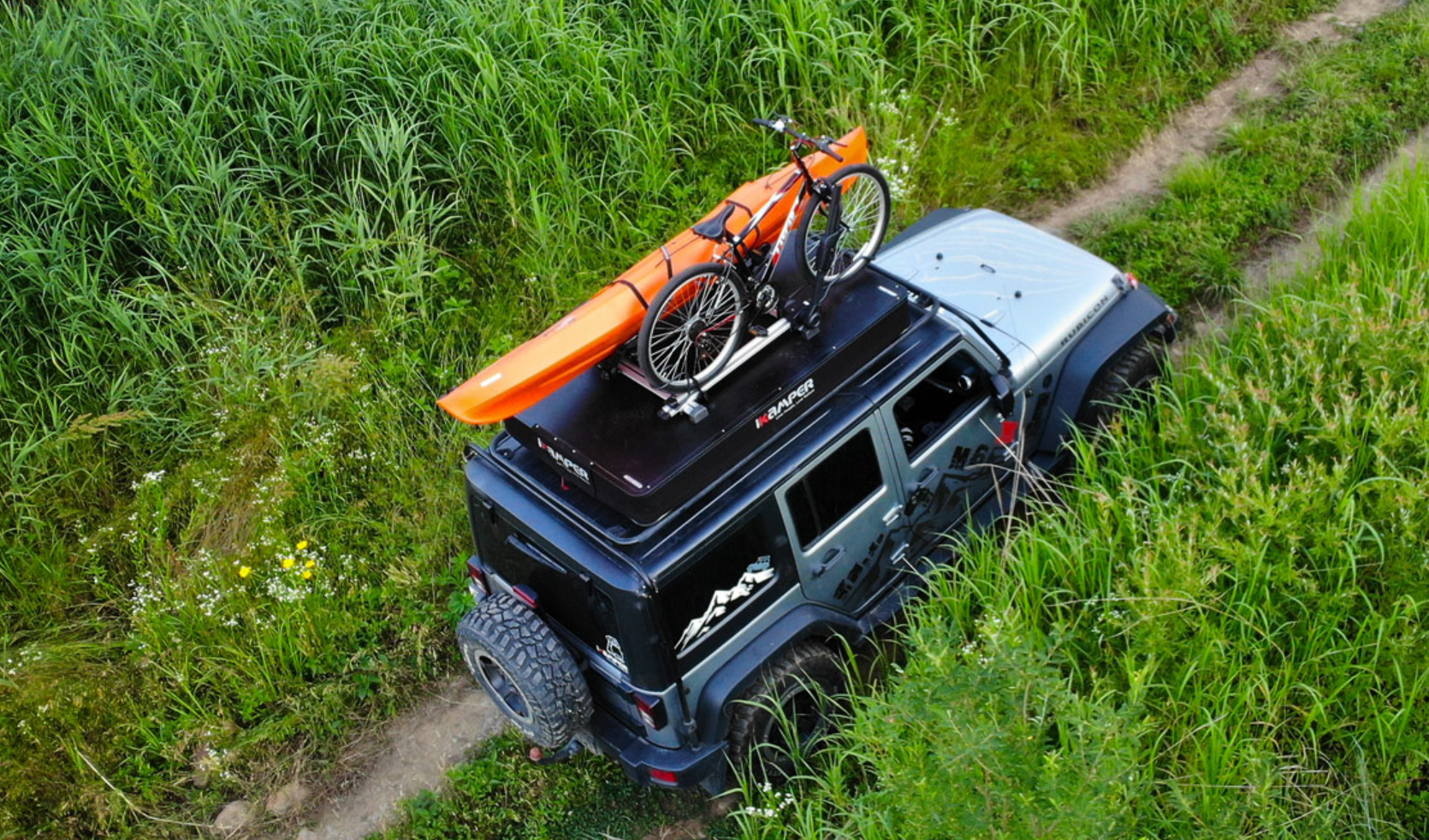 Un fuoristrada è parcheggiato su una strada sterrata. Sul tetto sono fissati una tenda da tetto rigida per 3 persone, una bicicletta e una canoa.