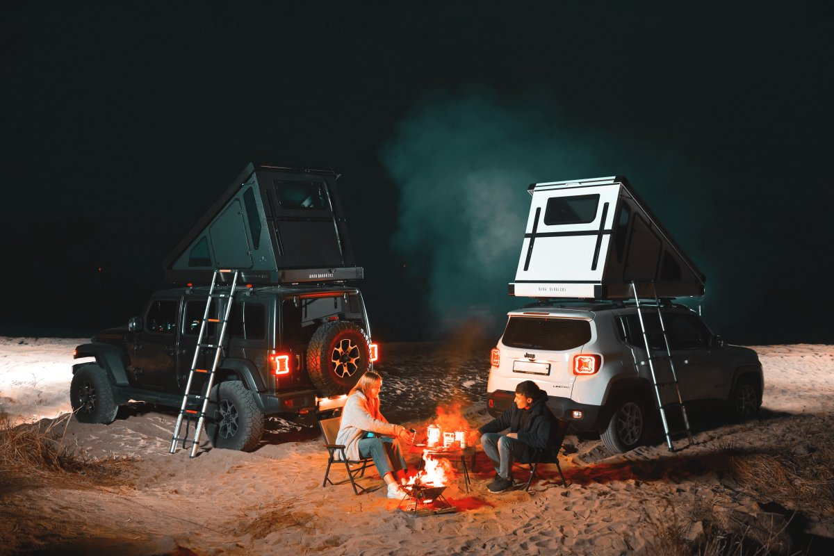Due persone sedute su sedie da campeggio intorno a un falò. Sullo sfondo le loro auto con tende da tetto rigide Dark Barracks