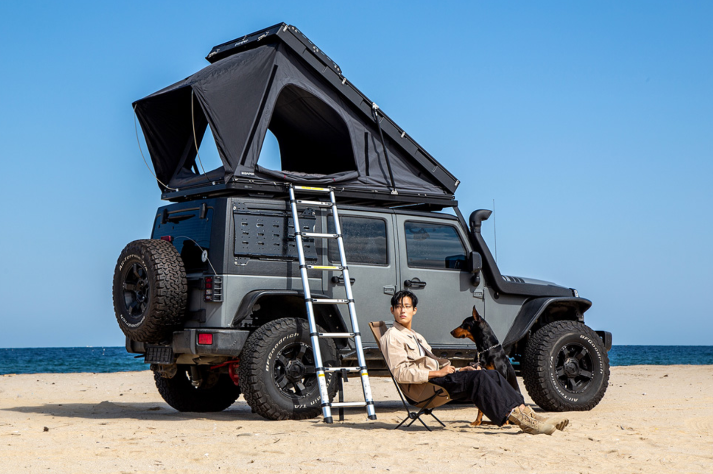 Uomo seduto su una sedia da campeggio con la mano sulla spiaggia, sullo sfondo la sua auto parcheggiata con tenda da tetto BDV Solo